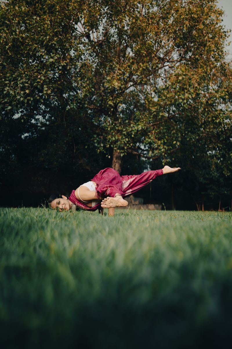 Shikha in an arm balance on the grass, under a large tree